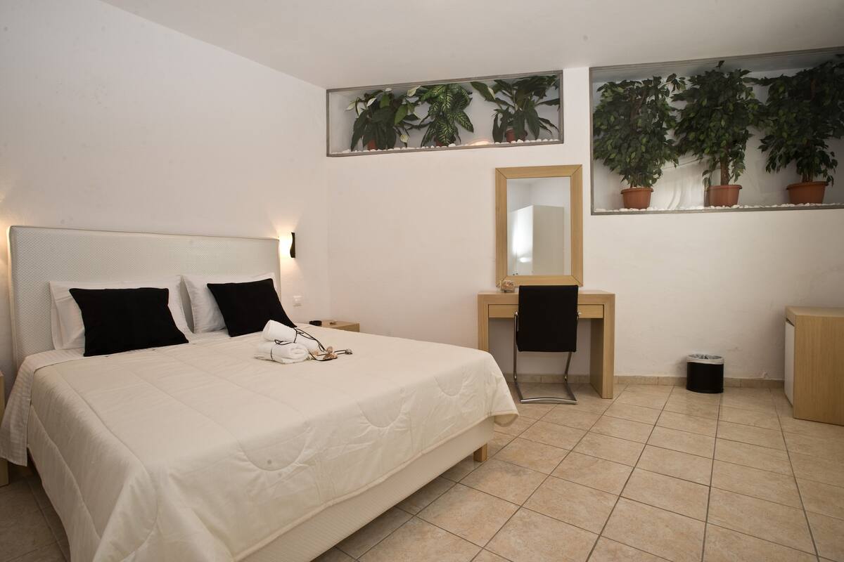 A bedroom with a bed, a desk, a chair, a mirror, and potted plants.