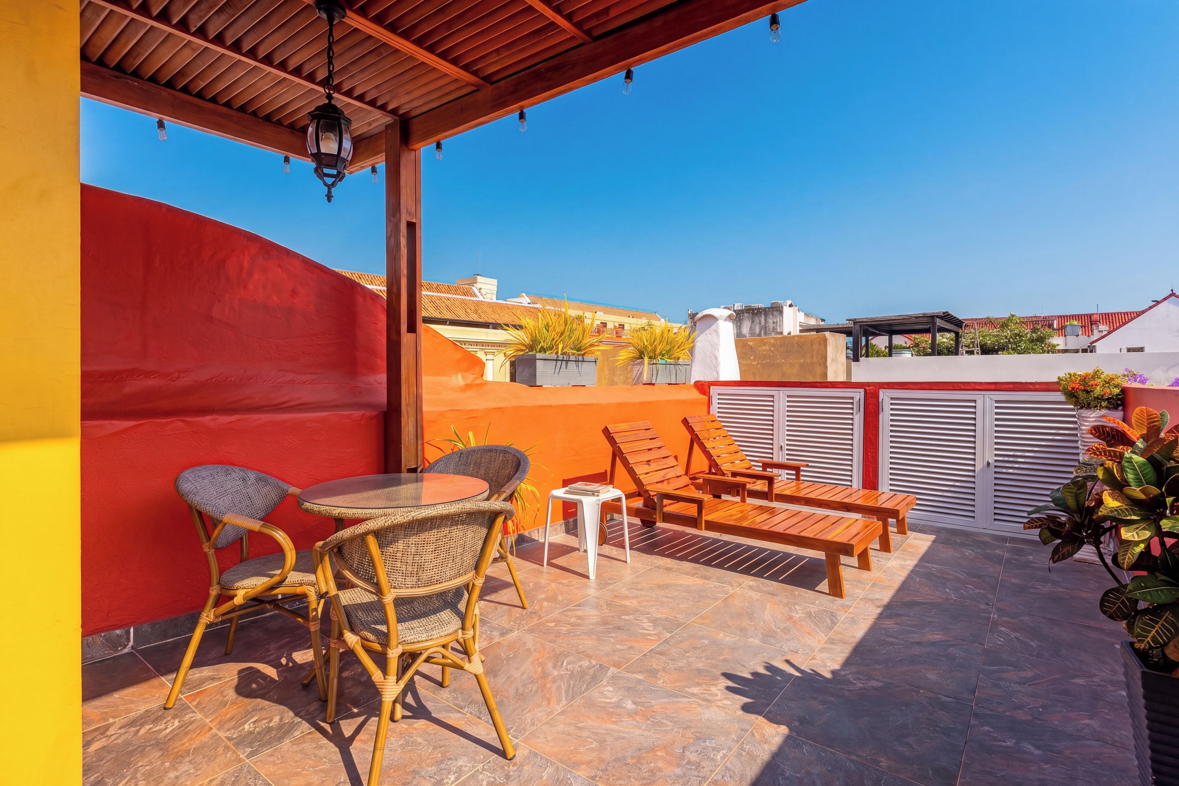 Un espace extérieur abrité, aménagé avec des chaises et une table en osier, un mur rouge et une vue sur le paysage urbain.