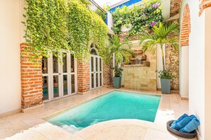 Piscina con pared de ladrillo y puertas de cristal, rodeada de plantas en macetas y una pequeña cascada.