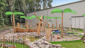 A zoo enclosure with a water feature, wooden structures, and a large building in the background. - Dünenvilla Superior (Lübeck)