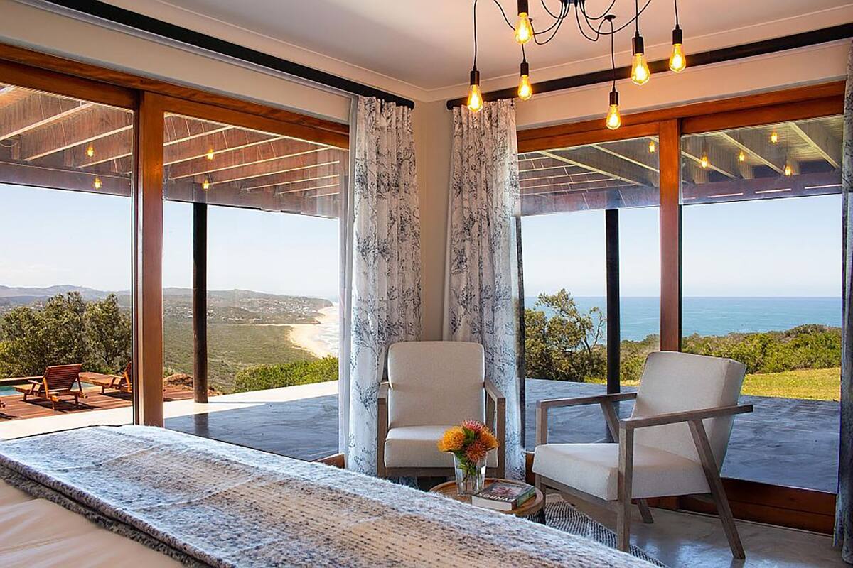 Une chambre moderne avec de grandes fenêtres, un lit, deux chaises et une vue sur l’océan.