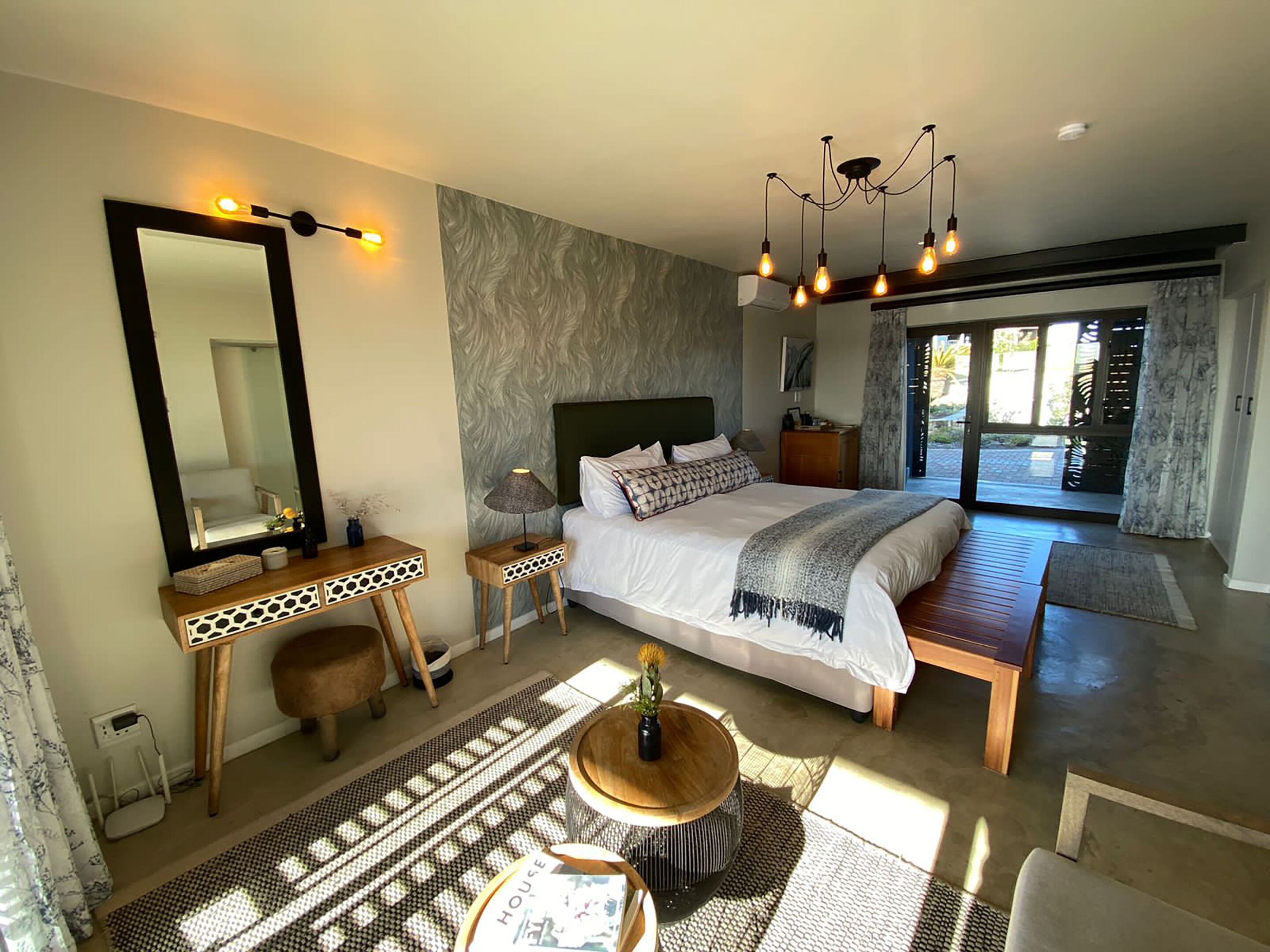 A modern bedroom with a large bed, a bedside table, a mirror, a lamp, and a view of the outdoors.
