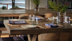 Une table à manger dressée avec des assiettes, des verres et des couverts, offrant une vue sur une étendue d’eau et des montagnes en arrière-plan.