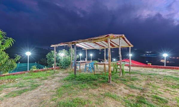 An outdoor seating area with tables and chairs, surrounded by greenery and a thatched-roof structure. - Treebo Om Villa With Mountain View (Dhari)
