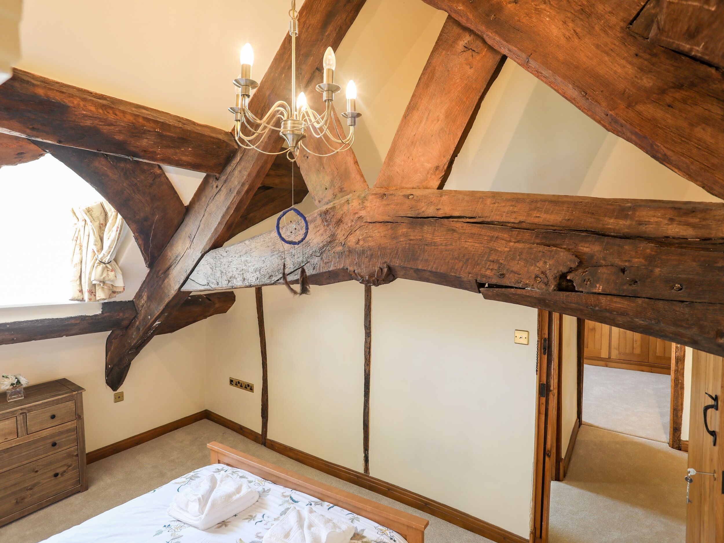 A room with exposed wooden beams, a chandelier, a bed with a floral bedspread, and a dresser with a small decorative item.