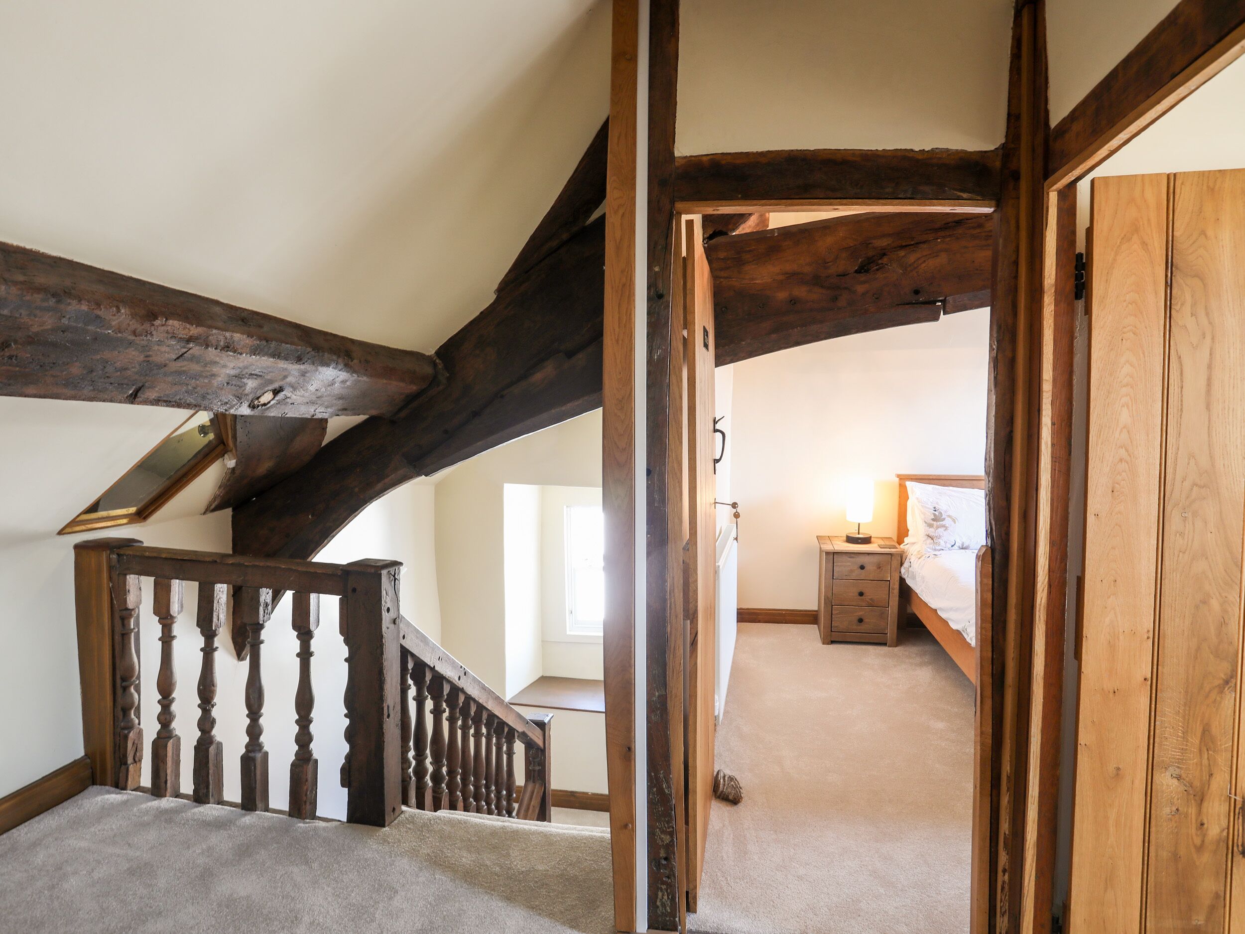 A staircase with wooden beams and a carpeted floor leading to a bedroom with a bed and a bedside table.