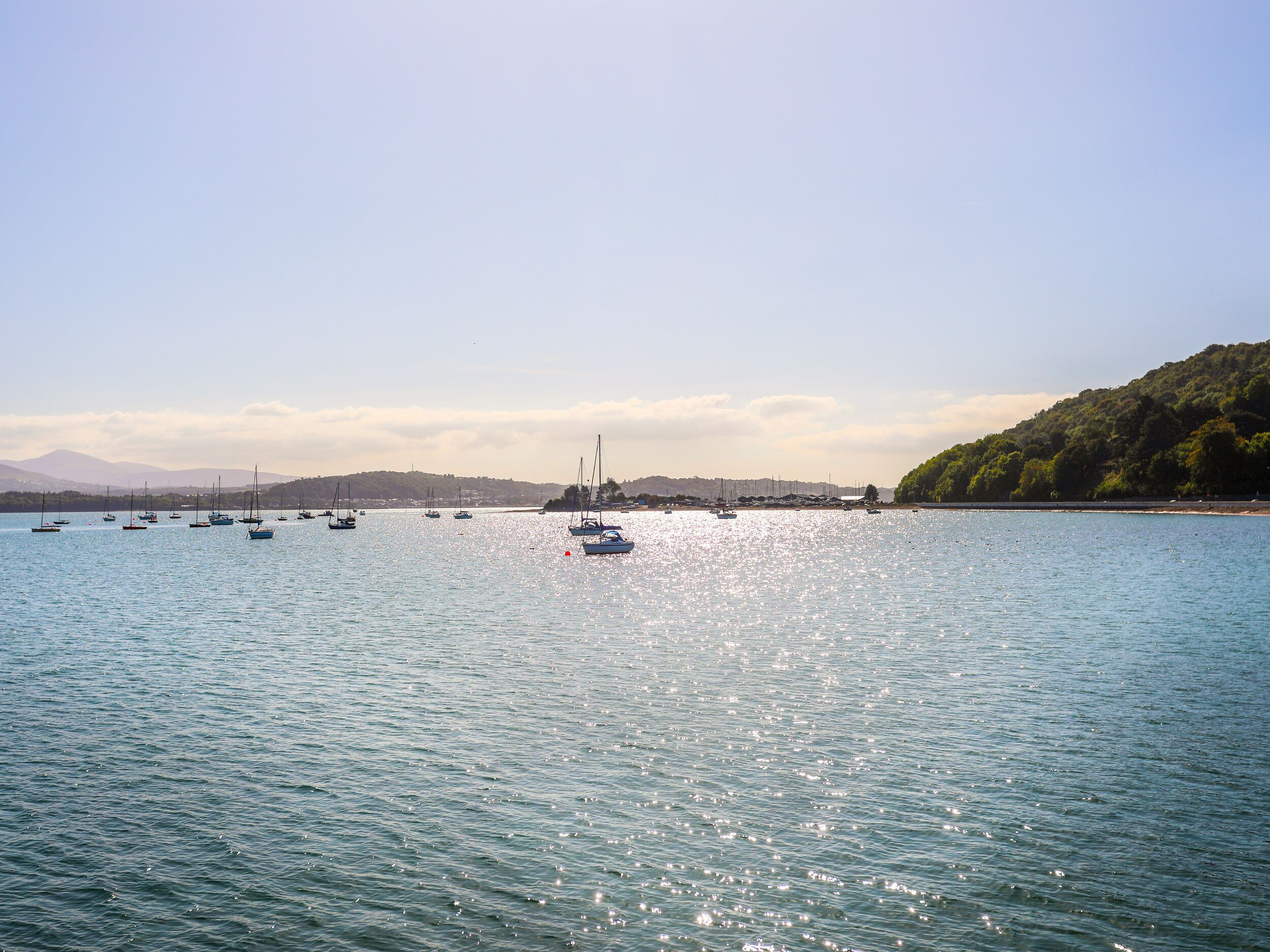 A calm lake with several sailboats anchored, surrounded by a hilly landscape and clear skies.