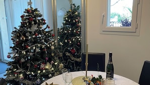 Una mesa de comedor dispuesta con platos dorados, copas y una botella de vino, rodeada de árboles de Navidad y un espejo.