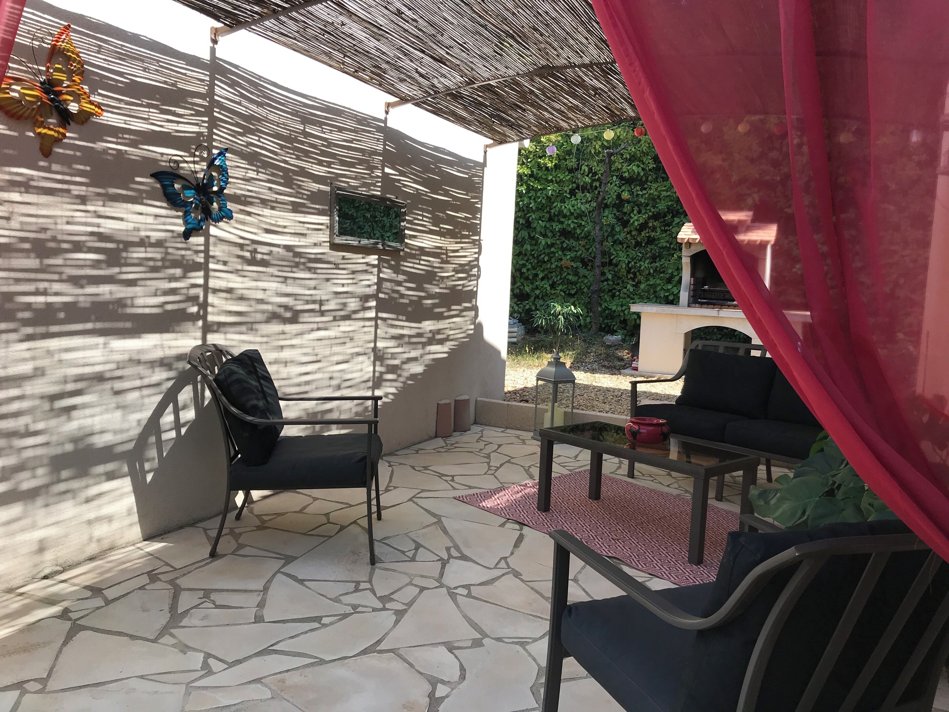 Un espace patio avec un sol en pierre, deux chaises, une petite table, un papillon décoratif et un téléviseur fixé au mur.