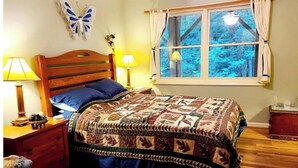 A bedroom with a wooden bed, two bedside lamps, a window with curtains, and a decorative butterfly on the wall. - Spacious suite surrounded by nature with living room, fireplace, porch, fire pit (Blowing Rock)