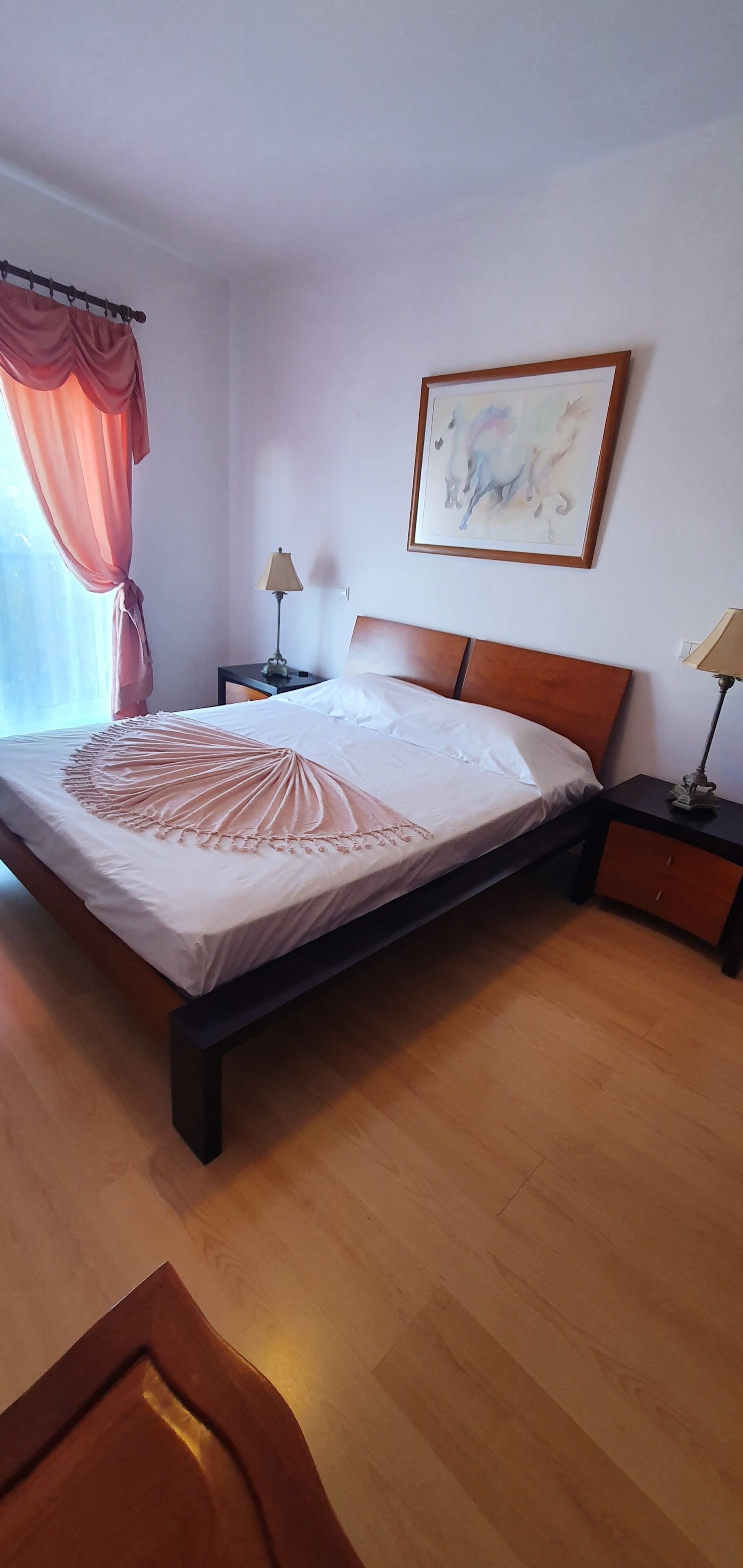 A neatly made bed with a decorative throw, a bedside table with a lamp, a framed abstract painting on the wall, and a window with curtains.