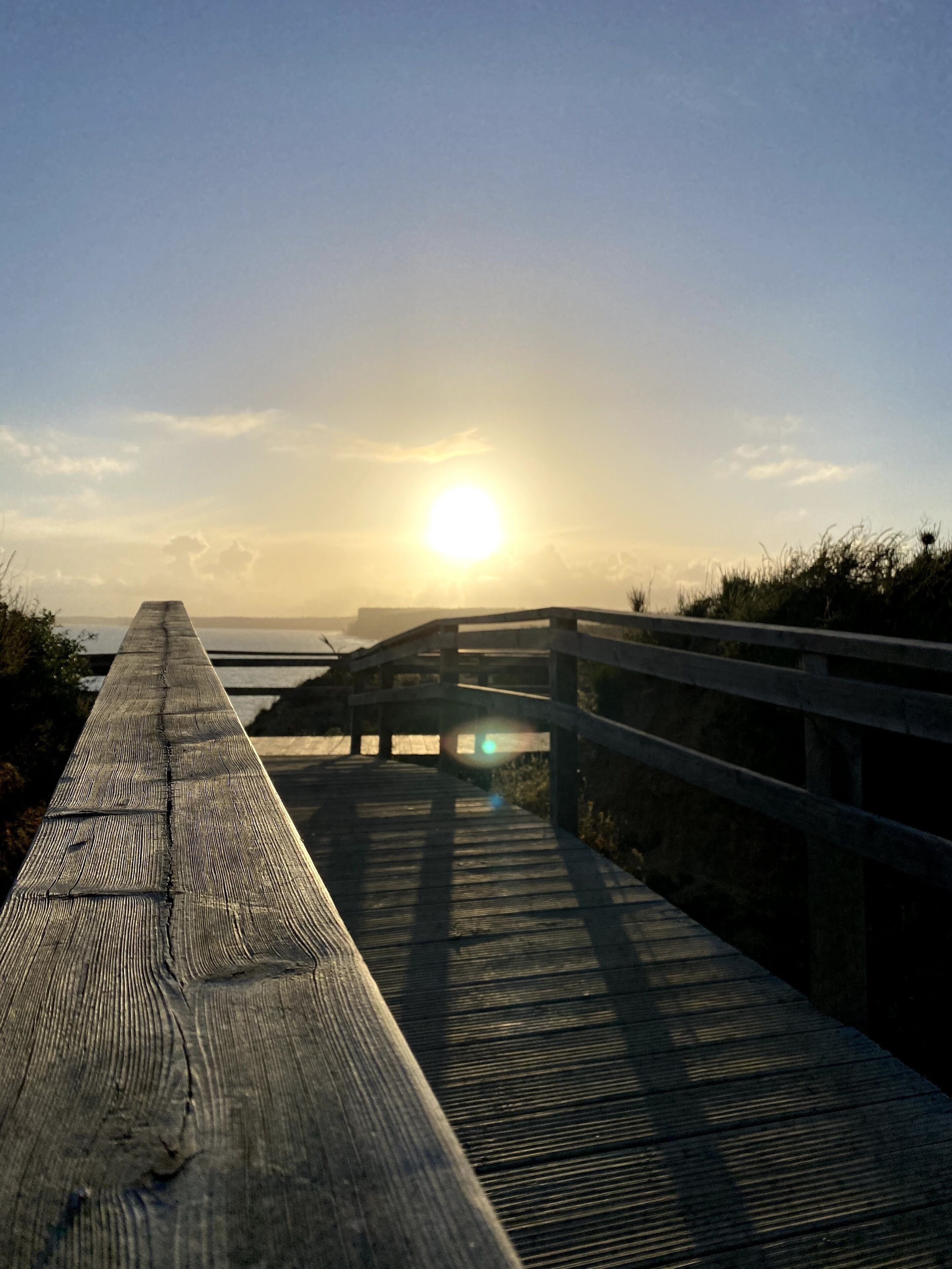 A wooden boardwalk leading to a bridge over water, with the sun setting in the background.