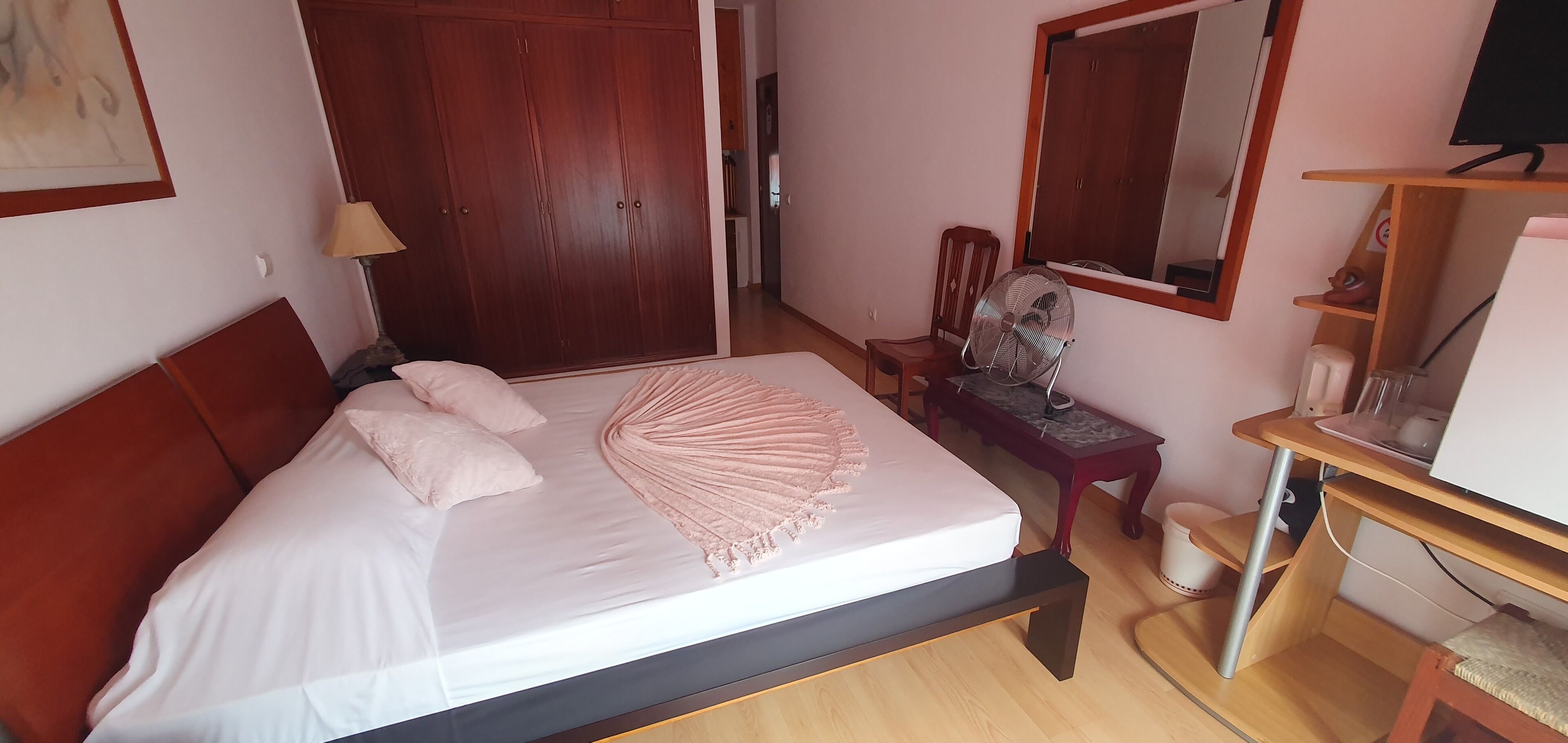 A neatly made bed with a pink throw, a wooden wardrobe, a chair, a desk with a fan, and a mirror.