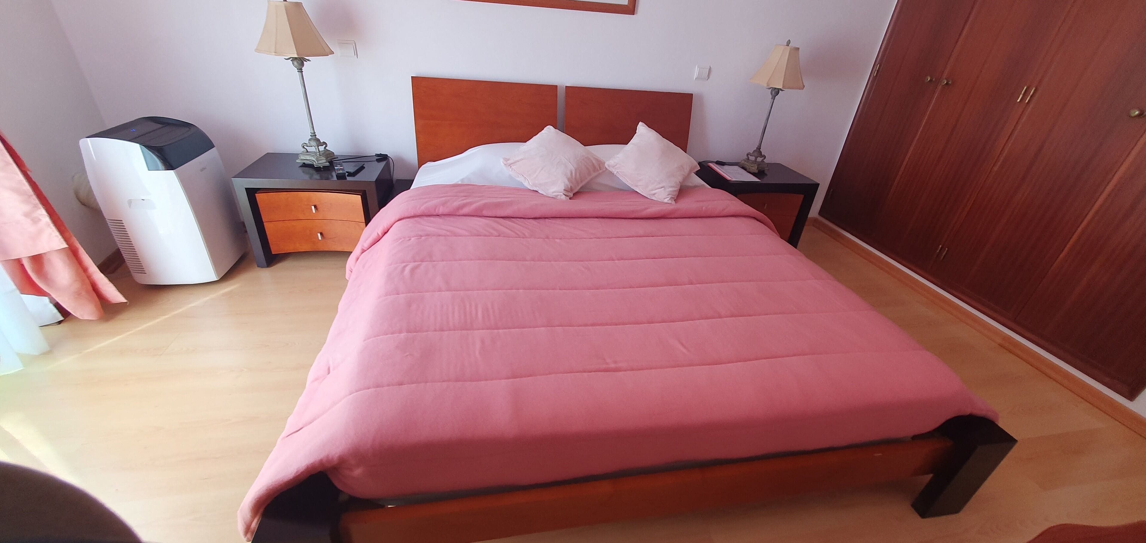 A neatly made bed with a pink bedspread, two bedside tables with lamps, a wooden wardrobe, and a white air conditioner.