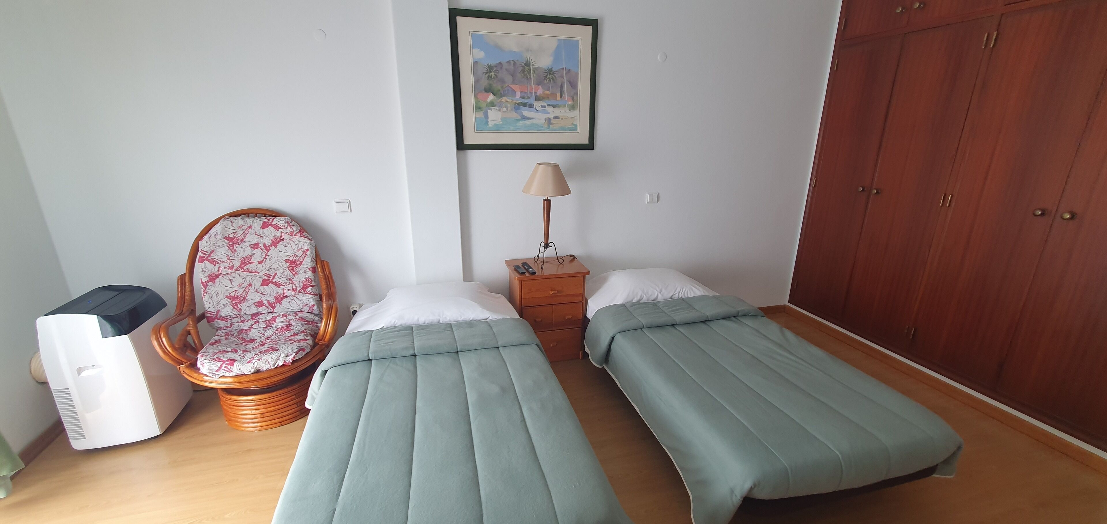 A room with two single beds, a wicker chair, a bedside table with a lamp, a heater, and a wardrobe.