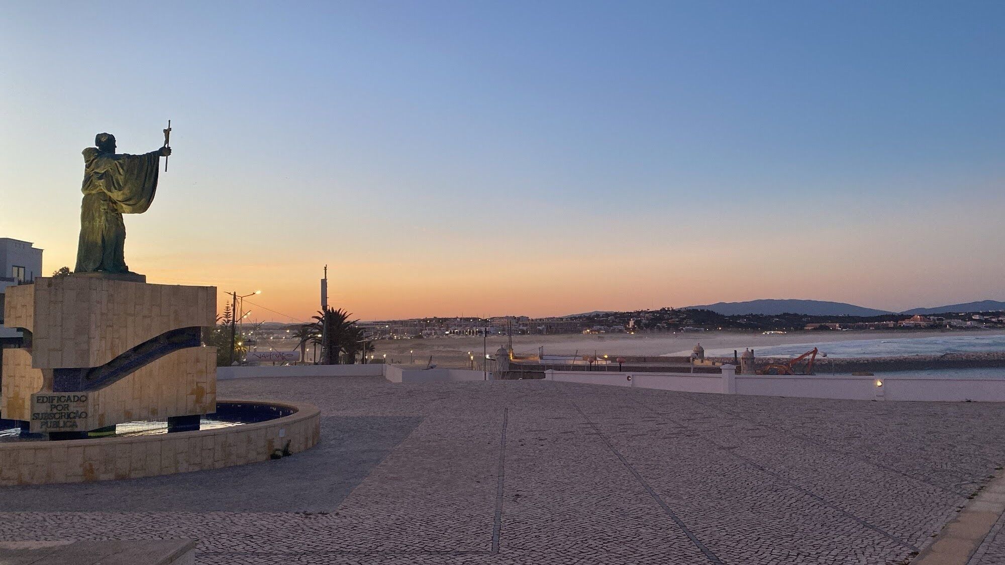 A statue on a pedestal in a public square with a view of the sea and distant hills.