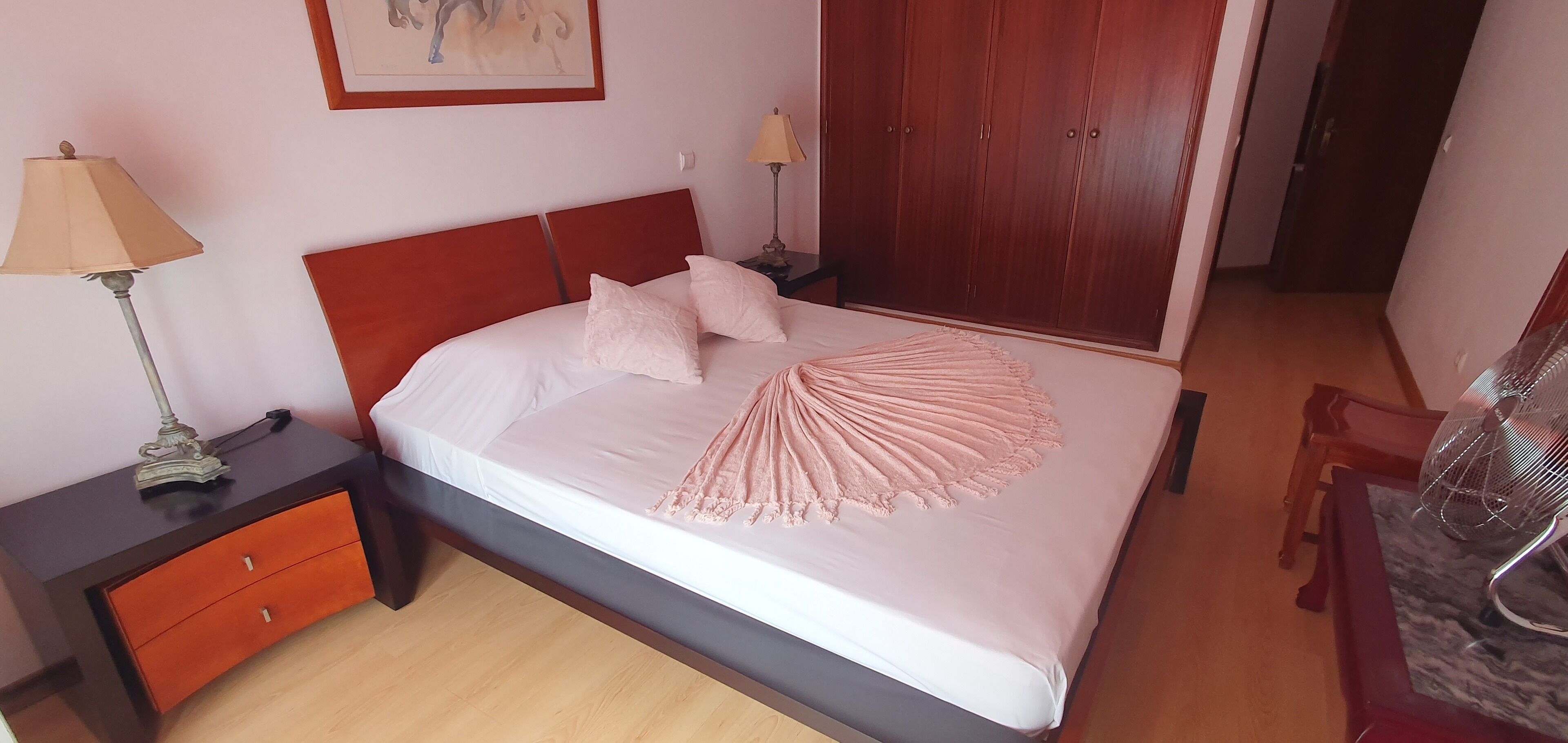 Un letto rifatto con cura, una coperta decorativa rosa, un comodino in legno con una lampada e un armadio in una camera d'albergo.