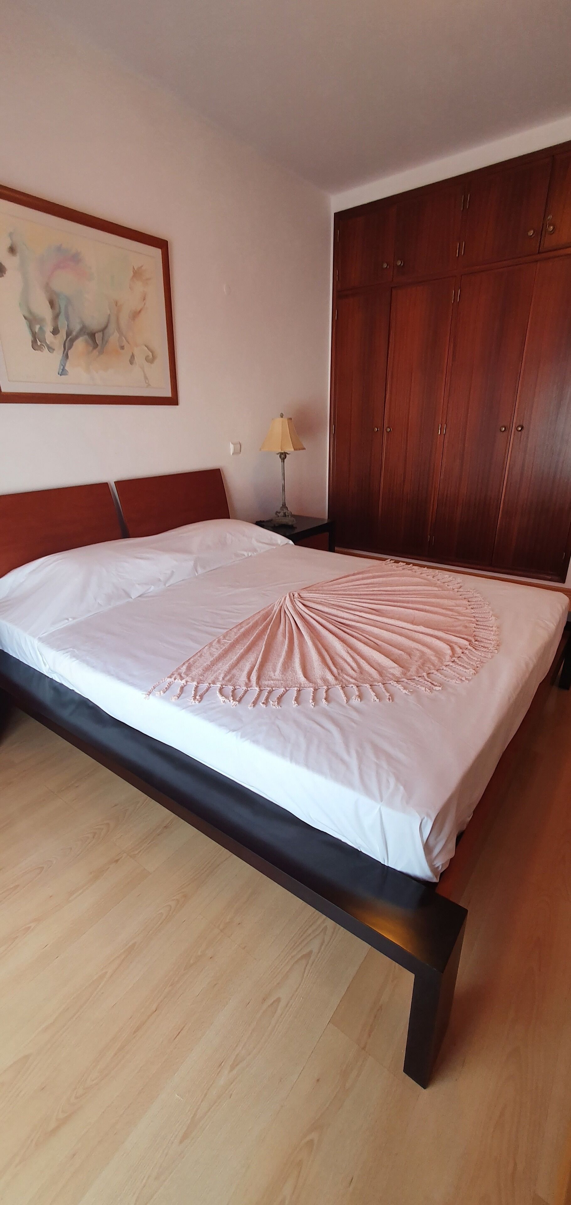 A bedroom with a bed, wooden floor, a wardrobe, a lamp, and a painting on the wall.