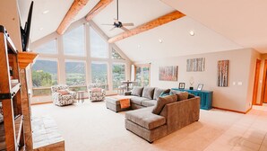 A spacious living room with a large gray sectional sofa, two patterned armchairs, and a coffee table. The room features a high ceiling with exposed wooden beams, large windows offering a view of the outdoors, and a ceiling fan. - Beautiful 30+ Stay | Amazing Views | Large Deck | Air Conditioning (Ridgway)