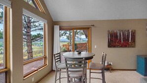 A dining area with a round table and four chairs, large windows with a view of trees and hills, and a painting on the wall. - Beautiful 30+ Stay | Amazing Views | Large Deck | Air Conditioning (Ridgway)