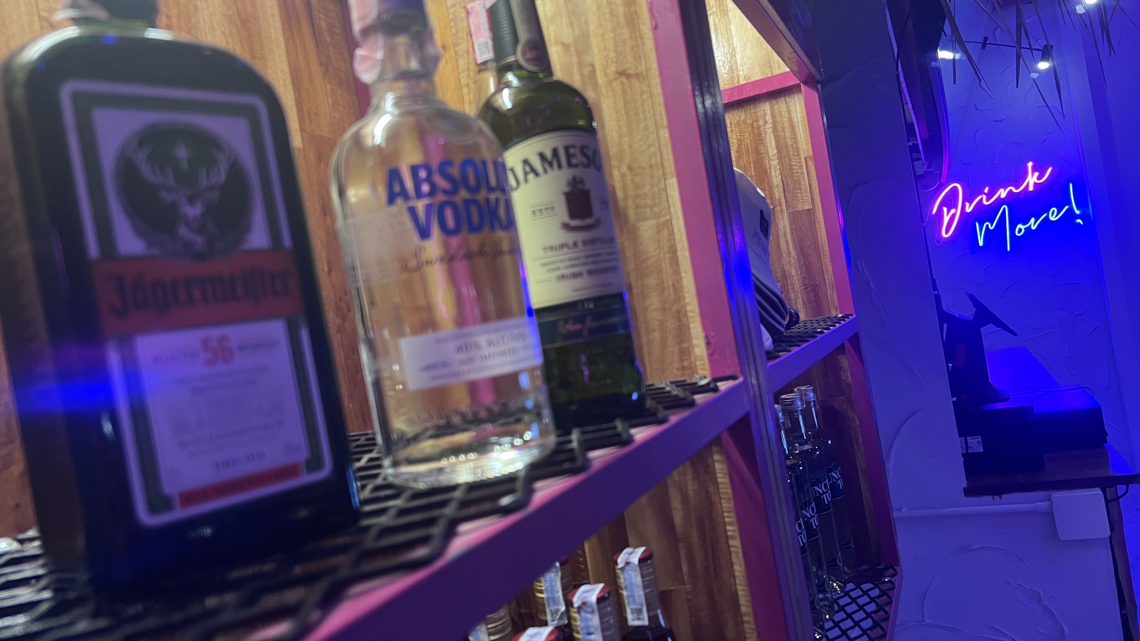 Angolo bar con bottiglie di Absolut Vodka e Jameson sugli scaffali, un neon con la scritta “Drink More!” e una lavagna per i menu.