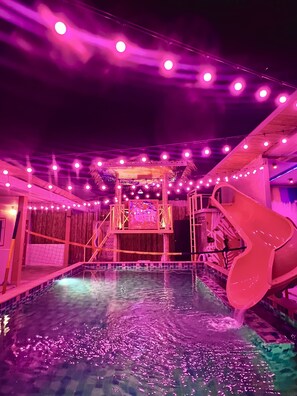 A water slide with a sign that reads "SLUMBER PARTY" in a pool area with string lights.