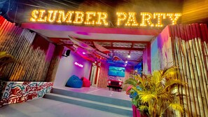 A vibrant entrance with a neon sign reading "SLUMBER PARTY" and a colorful, eclectic interior with a large screen and a blue bean bag chair.