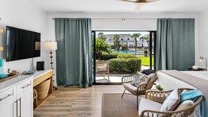 Une chambre d’hôtel moderne dotée d’une télévision à écran plat, d’un bureau avec une lampe, de meubles en osier et offrant une vue sur une piscine et la verdure environnante.