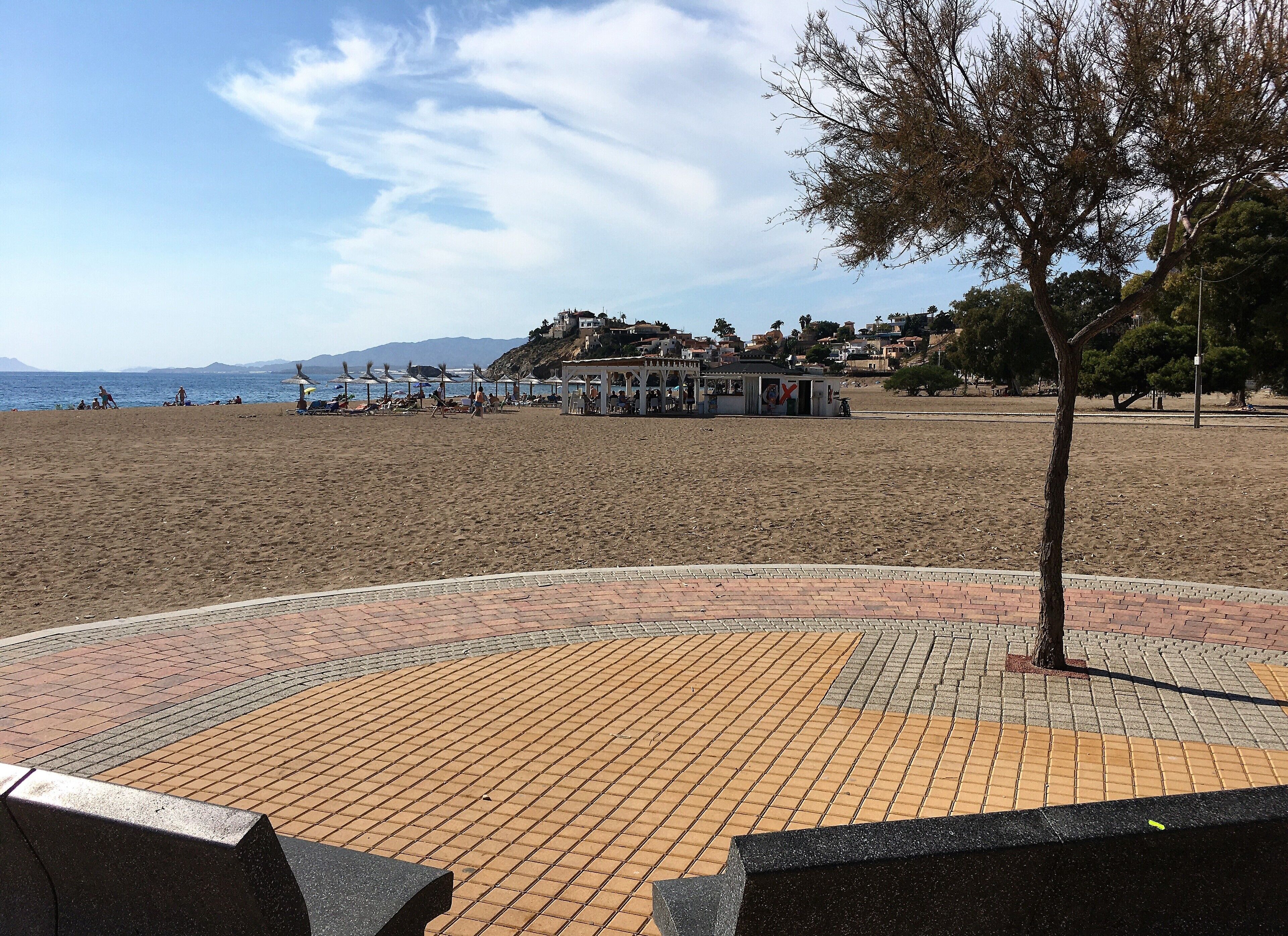 A beach with a paved walkway, umbrellas, and a building with a red 'X' sign.