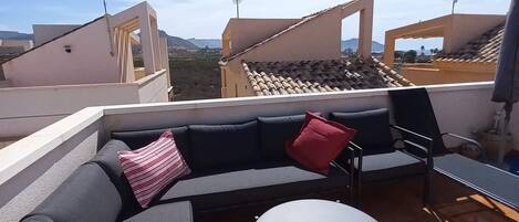 A rooftop terrace with a sectional sofa, a round table, and a view of the city and mountains in the background.