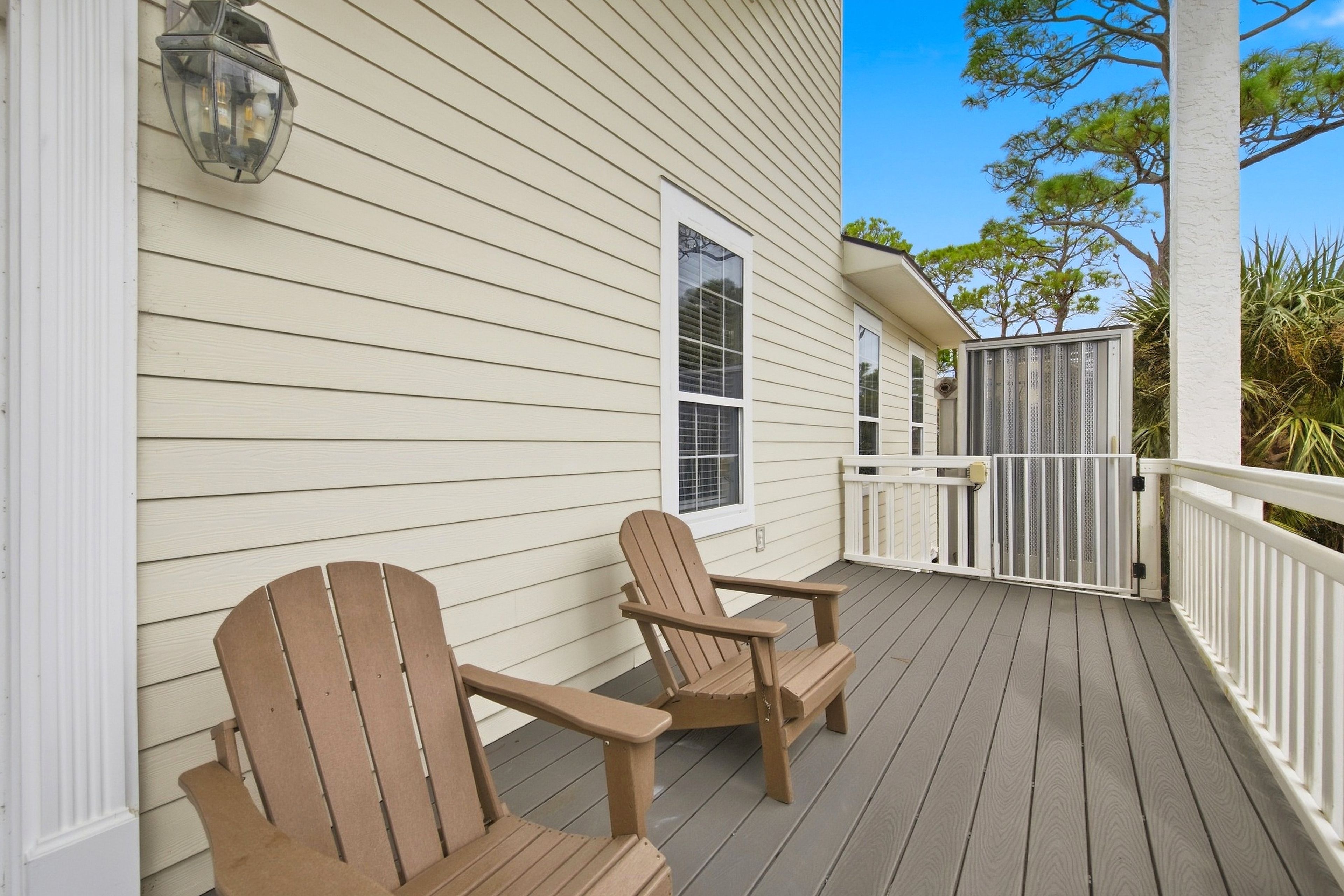 A wooden deck with two Adirondack chairs, a white railing, and a house with a window and a door.