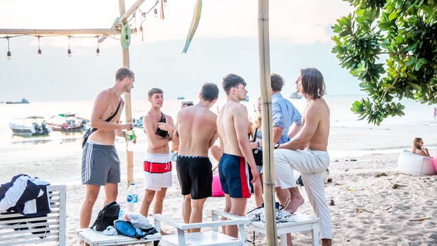 Un grupo de personas en una playa con una estructura de madera sobre ellas, un banco y una mesa con objetos encima.