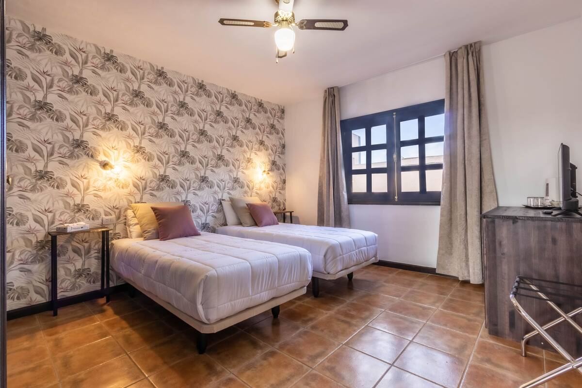 A room with two beds, a ceiling fan, a window with curtains, and a desk with a television.
