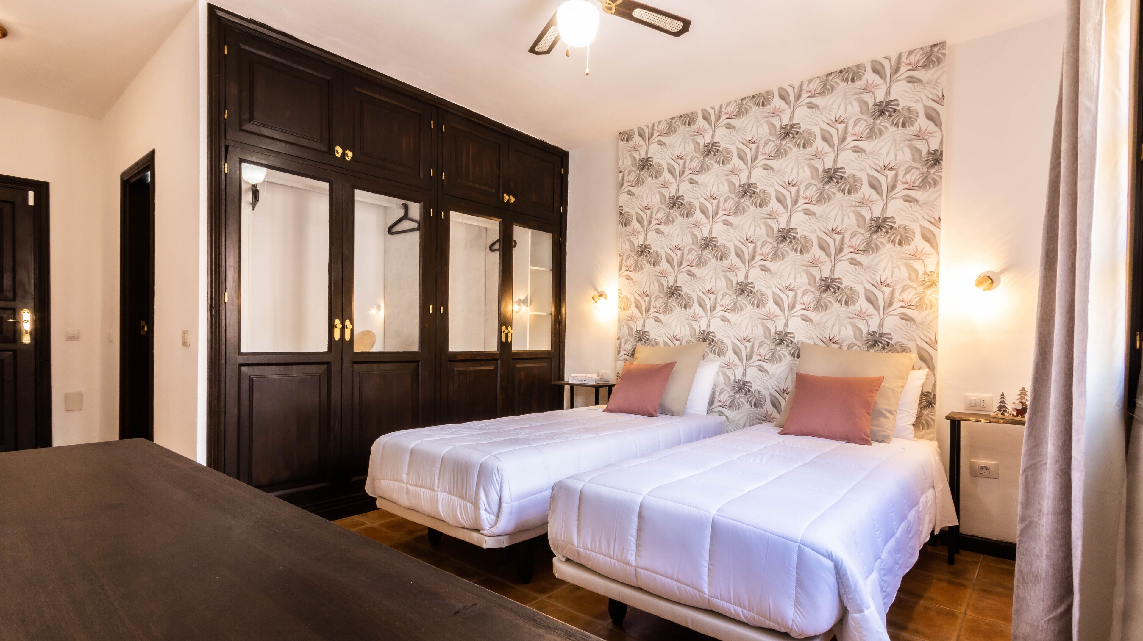 A hotel room with two beds, dark wooden wardrobe, floral wallpaper, and bedside lamps.