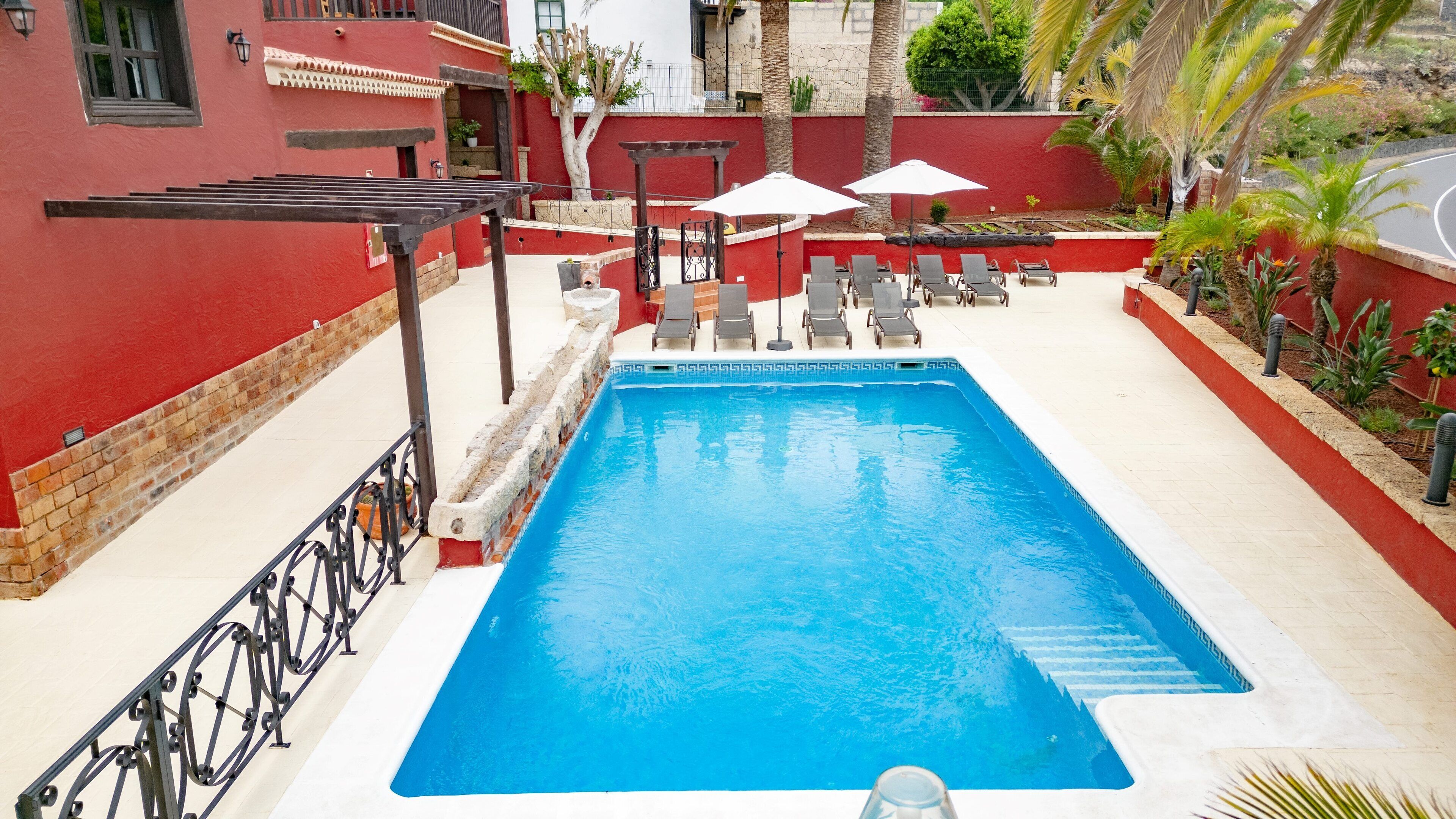 Una piscina rectangular con superficie de agua azul cristalina, rodeada por un muro rojo y una solana con baldosas blancas. Hay sillones de descanso y sombrillas a lo largo de la piscina.