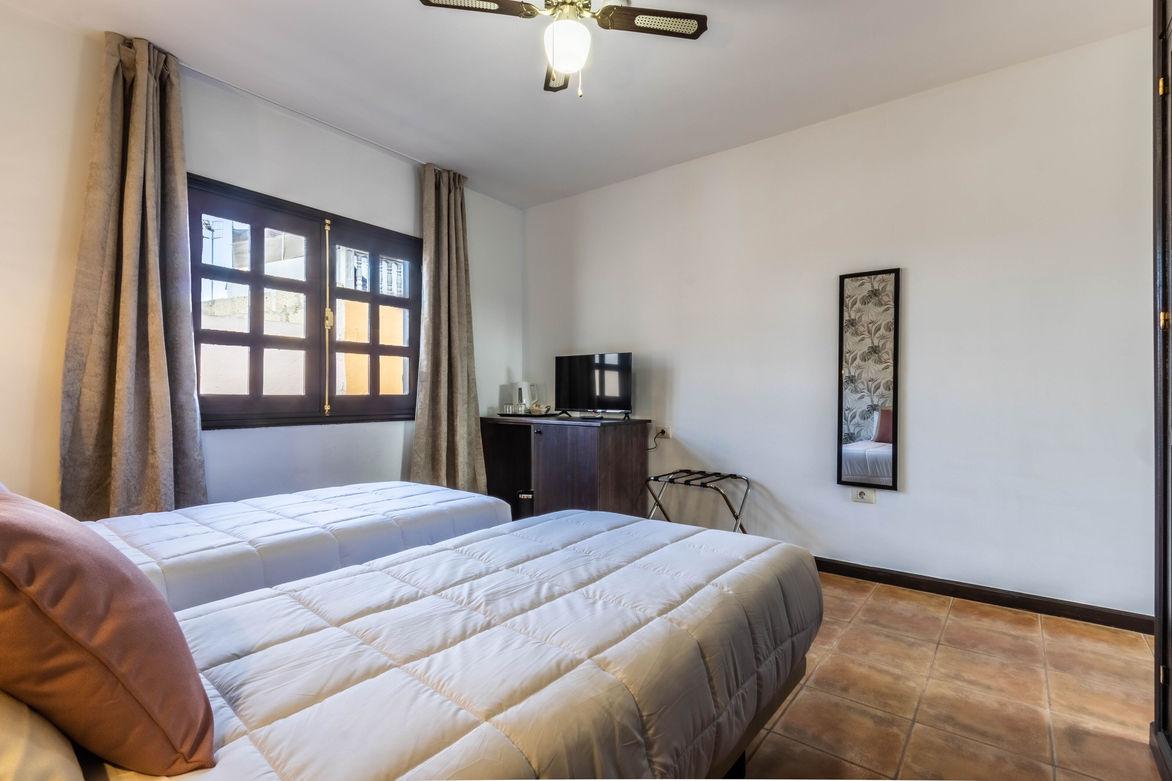 A hotel room with two beds, a ceiling fan, a TV, and a window with curtains.