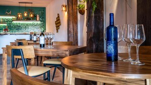 Interior de restaurante con mesas y sillas de madera, zona de bar con estanterías llenas de botellas y una planta grande en maceta.