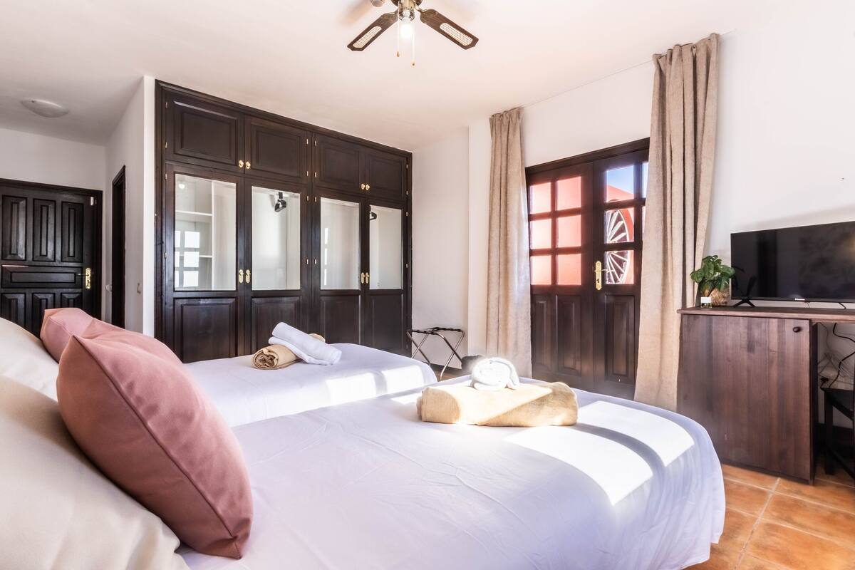 A bedroom with a large bed, dark wooden wardrobe, a desk with a television, and a window with curtains.