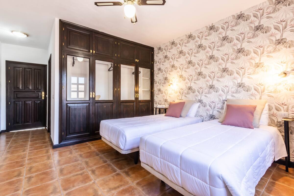 A bedroom with two beds, a wardrobe, a ceiling fan, and a floral wallpaper.