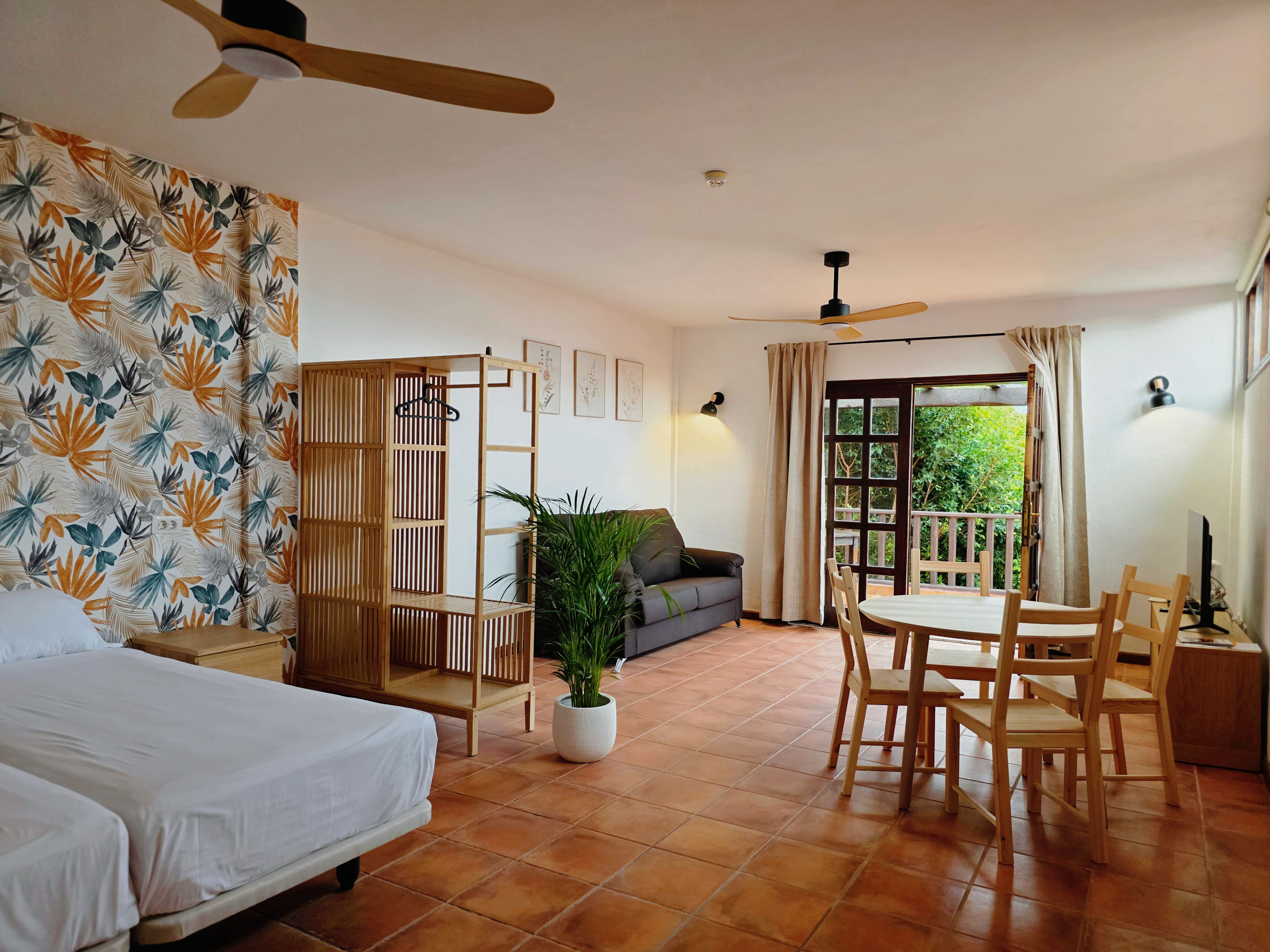 A hotel room with a bed, a seating area with a table and chairs, a potted plant, and a balcony with a view.