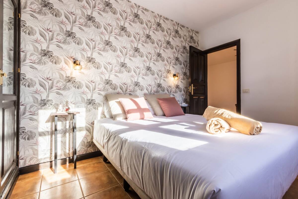 A bedroom with a floral wallpaper, a bed with multiple pillows, a bedside table, and a nightstand.