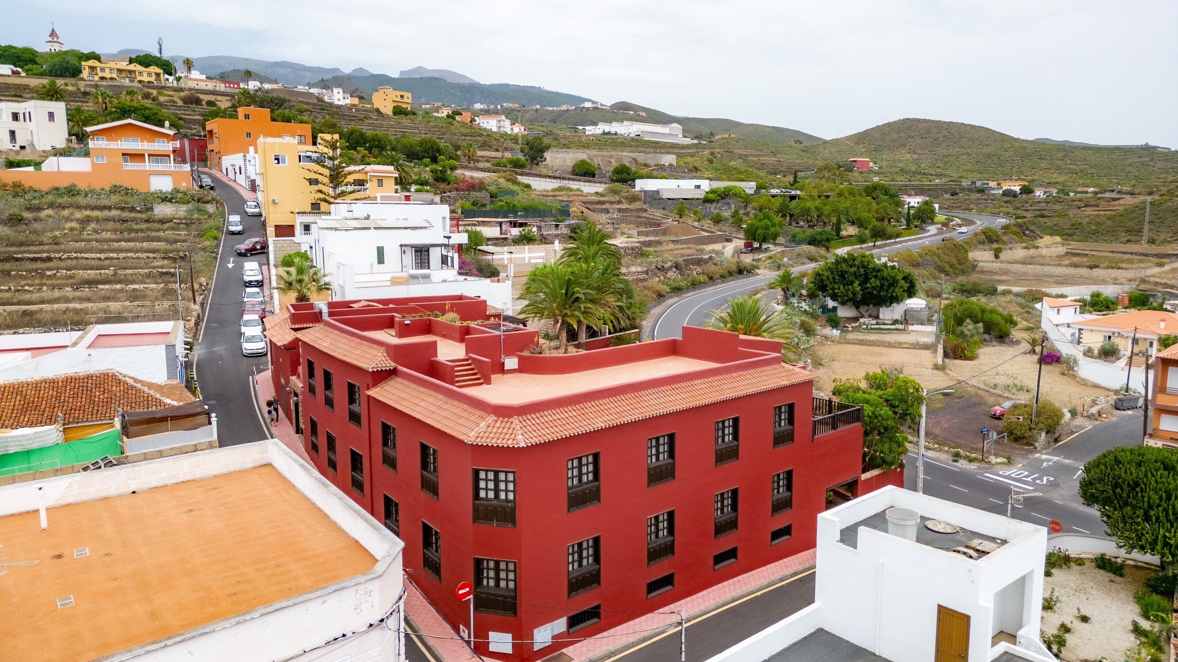 Un edificio rojo con varias ventanas y techo plano, rodeado por una calle con coches aparcados y algunos edificios al fondo.