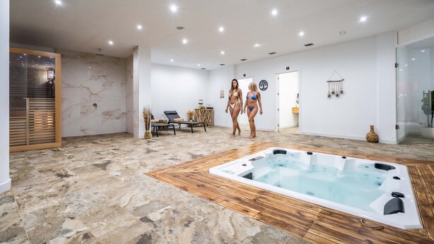 Un área moderna de piscina interior con solárium de madera, un jacuzzi y una zona de descanso con mesa y sillas.