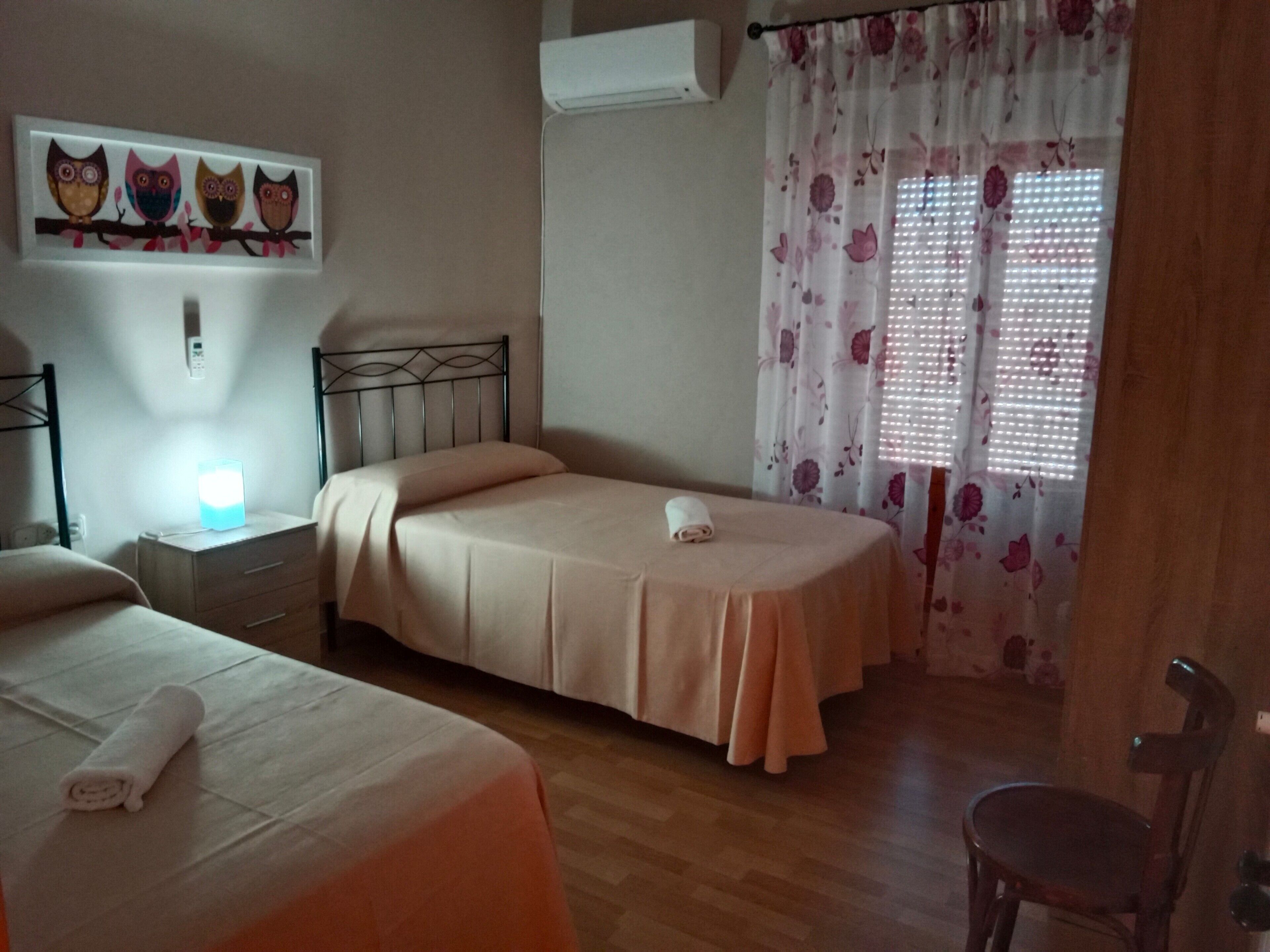 Un dormitori amb dos llits, una tauleta de nit, aire condicionat fixat a la paret i una finestra amb cortines florals.