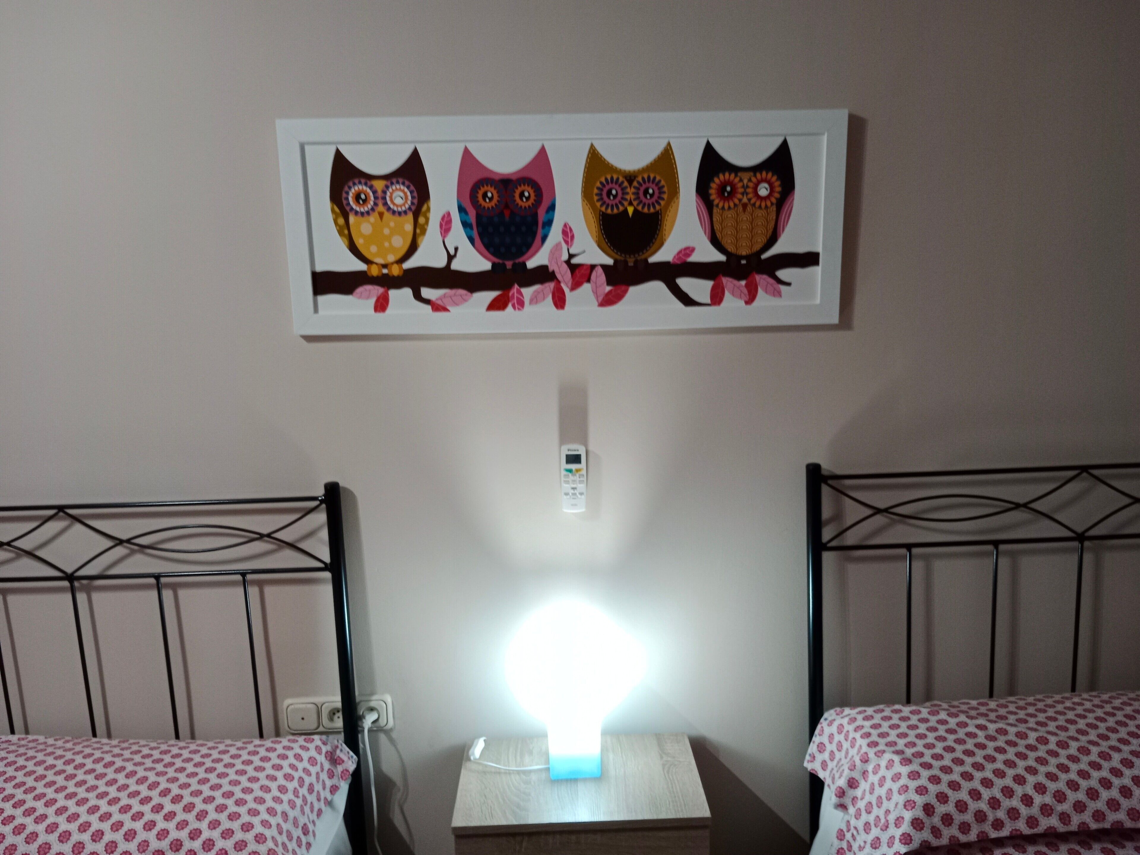 A room with two beds, a nightstand, a wall-mounted lamp, and a framed artwork of owls.