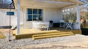 En hvid træhytte med en overdækket terrasse, et bord med stole og et grillområde.