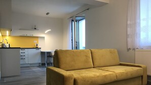 A modern living room with a yellow sofa, a kitchen area with white cabinets, and a large window providing natural light.