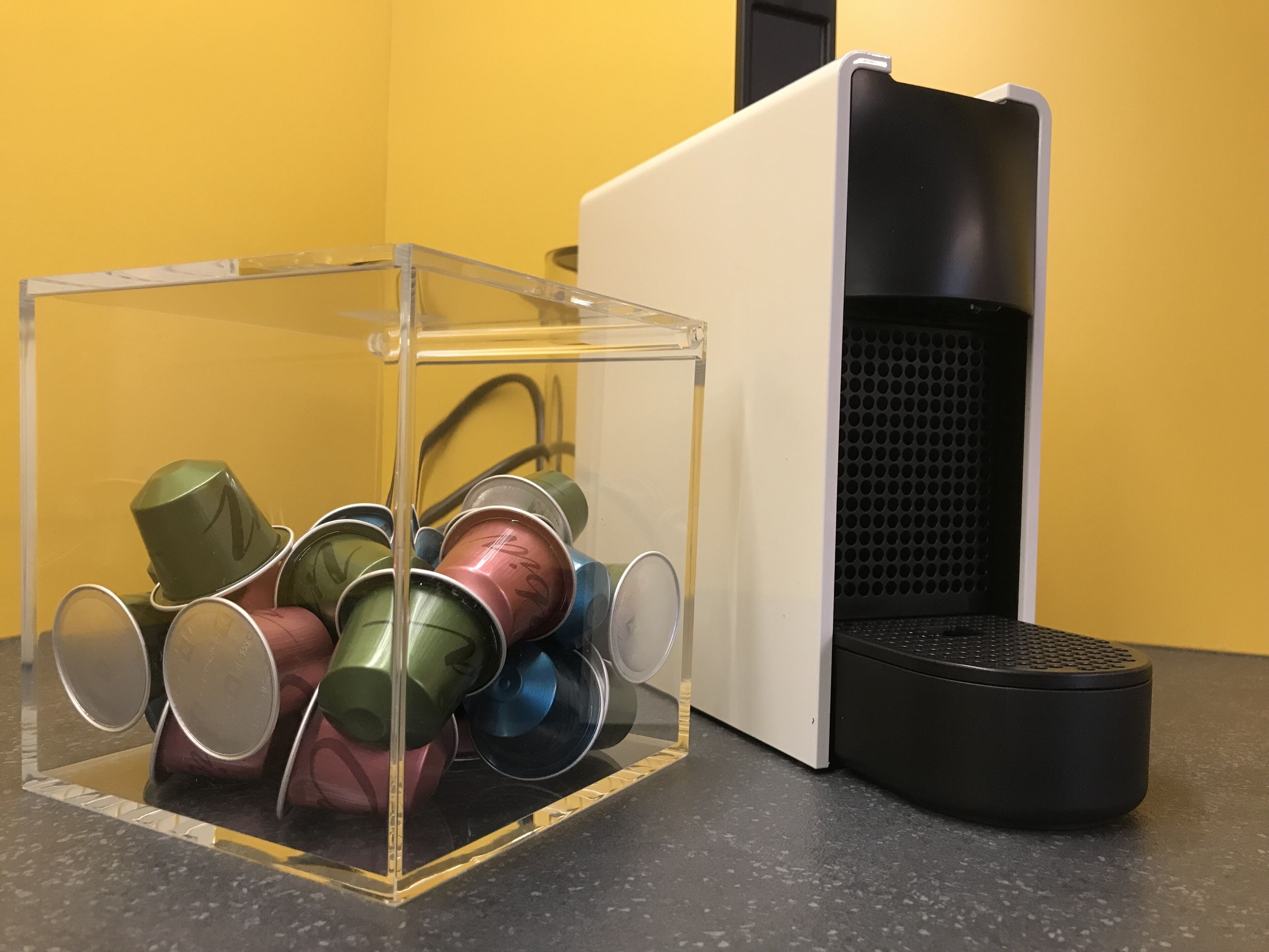 A clear plastic storage box with metal cups inside, placed on a black countertop next to a black and white speaker.