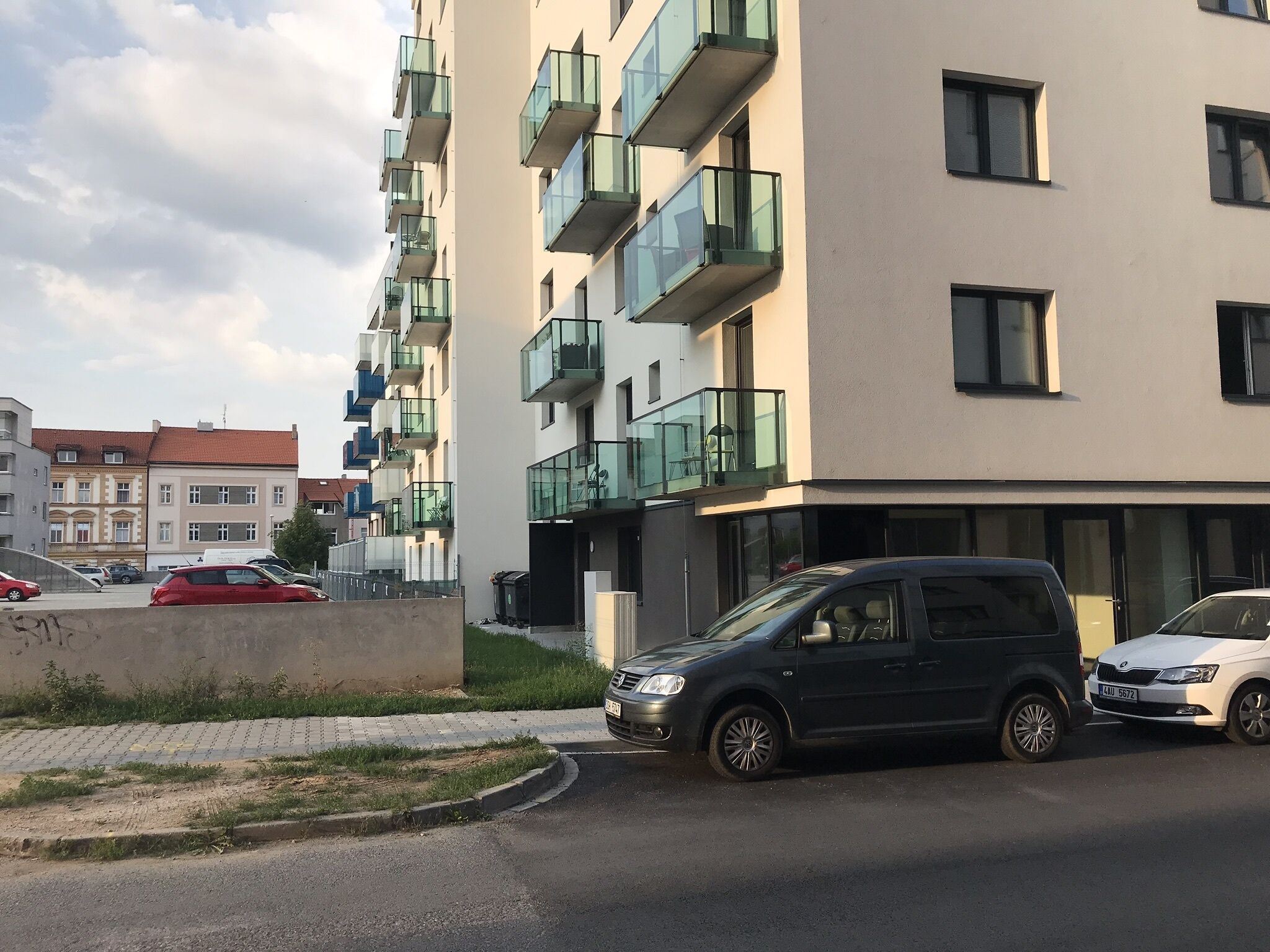 A modern apartment building with multiple balconies, a parking area, and a road with cars.