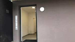 An entrance to a building with a black door marked with the number 18, a set of four control panels on the wall, and a circular light fixture above the door.